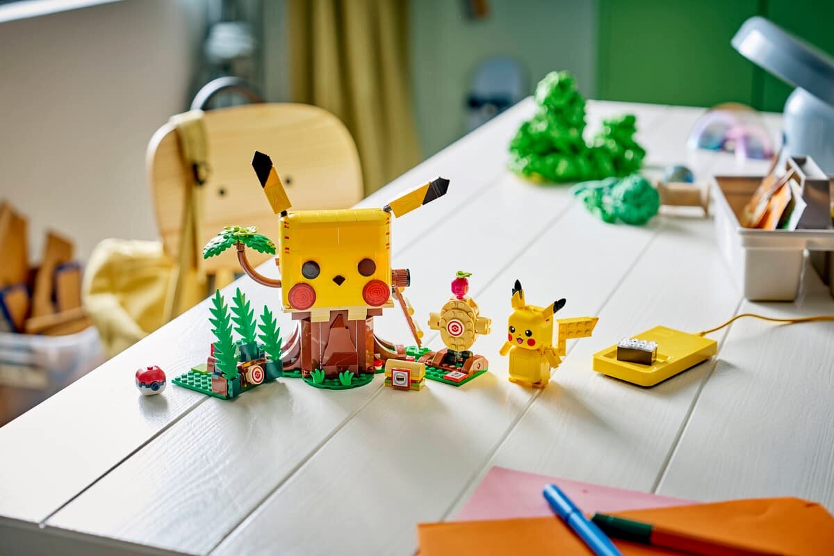 Na białym stole znajduje się zestaw klocków przedstawiający dom treningowy z postacią przypominającą Pikachu oraz kilka akcesoriów. W tle widać krzesło i różne przedmioty, w tym zieloną roślinę i pudełko z przyborami.