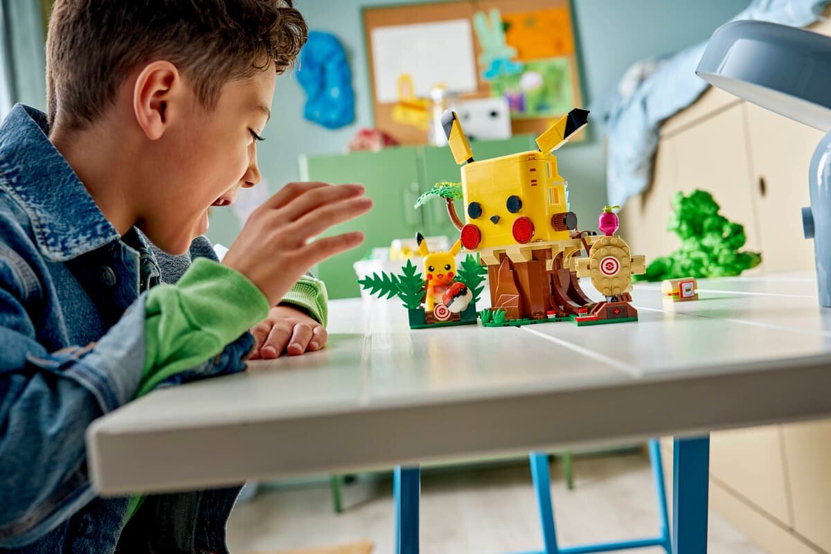 Na stole znajduje się zestaw LEGO z motywem Pikachu, przedstawiający dom treningowy z elementami roślinnymi i tarczami. W tle widać fragment pokoju z meblami i dekoracjami na ścianie.