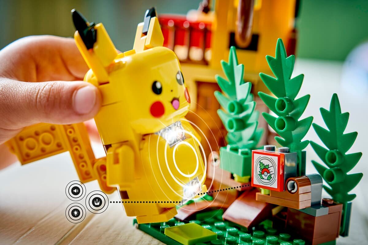 Na obrazie widać figurkę Pikachu z klocków LEGO, trzymaną w dłoni, obok której znajdują się zielone elementy przypominające rośliny oraz mały ekran z symbolem Poké Balla. W tle widoczna jest część konstrukcji z klocków LEGO.