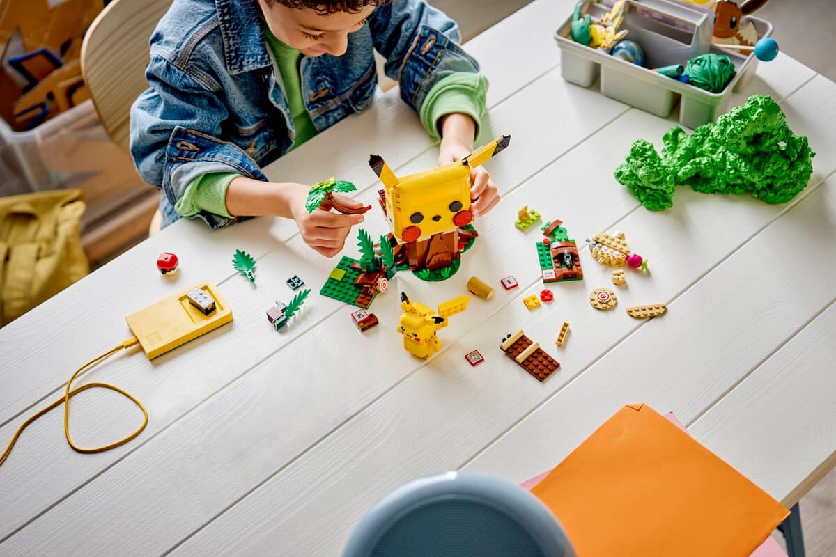 Na białym stole znajdują się klocki LEGO, w tym model Pikachu oraz inne elementy do budowy. Obok siedzi osoba, która bawi się klockami.