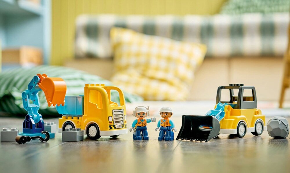Na obrazie znajdują się zabawkowe maszyny budowlane z klocków LEGO oraz dwie figurki pracowników. W tle widoczne są poduszki i rozmyte elementy wnętrza.