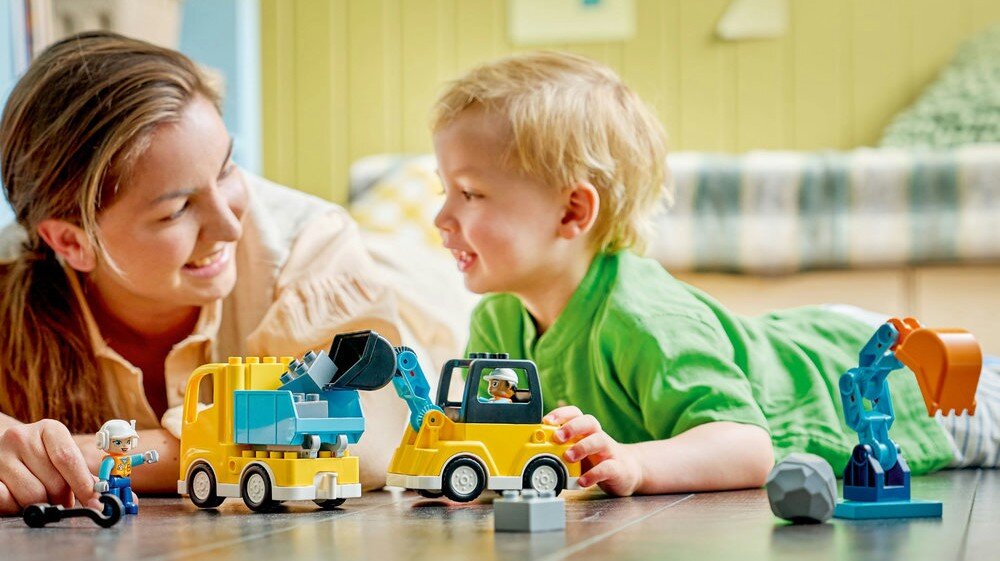 Dwoje ludzi bawi się zestawem klocków LEGO DUPLO, przedstawiającym maszyny budowlane. Na podłodze znajdują się kolorowe pojazdy i figurki.