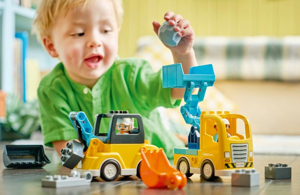 Dziecko bawi się zestawem klocków LEGO, przedstawiającym żółte maszyny budowlane z niebieskimi elementami. Na podłodze widoczne są różne części zestawu, w tym koparka i ciężarówka.