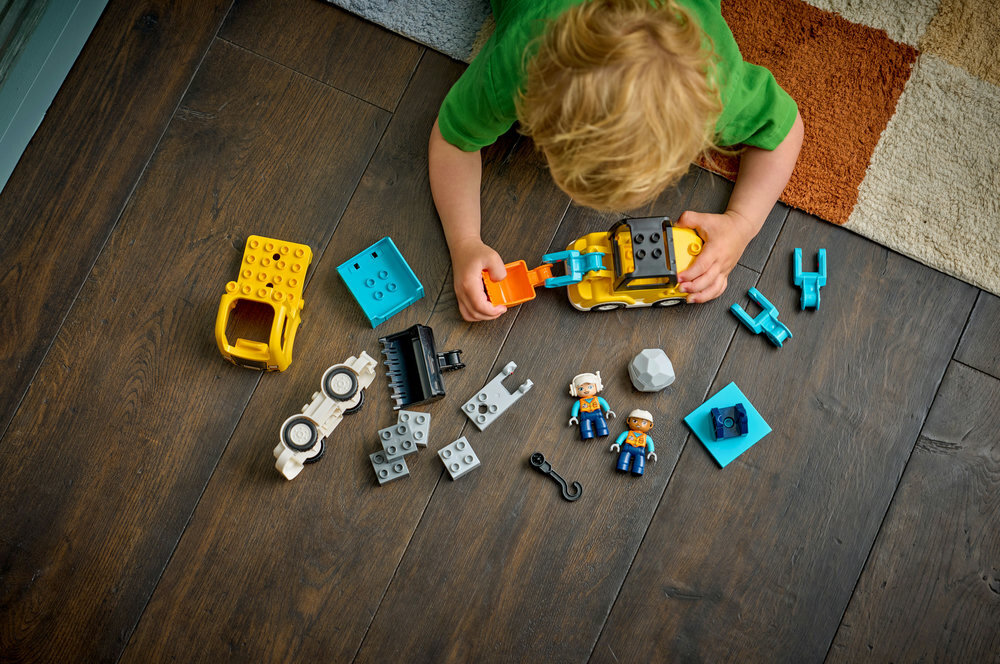 Dziecko bawi się zestawem klocków LEGO DUPLO, w tym elementami maszyn budowlanych i figurkami. Na podłodze rozłożone są różne części, takie jak koła, łyżka koparki i figurki robotników.