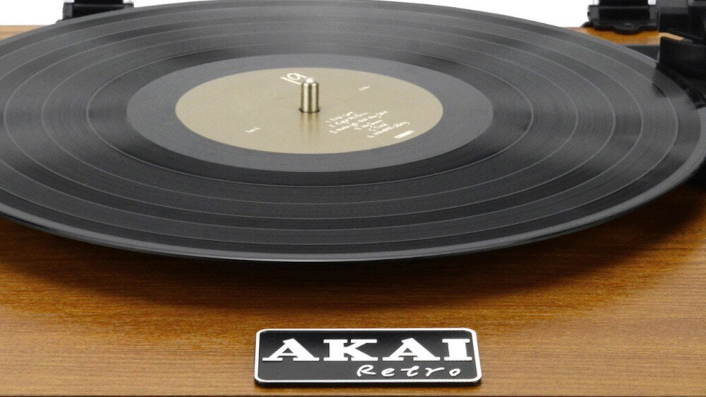 Na obrazie widoczny jest gramofon z czarną płytą winylową na drewnianej podstawie. Na podstawie znajduje się logo \'AKAI Retro\'.