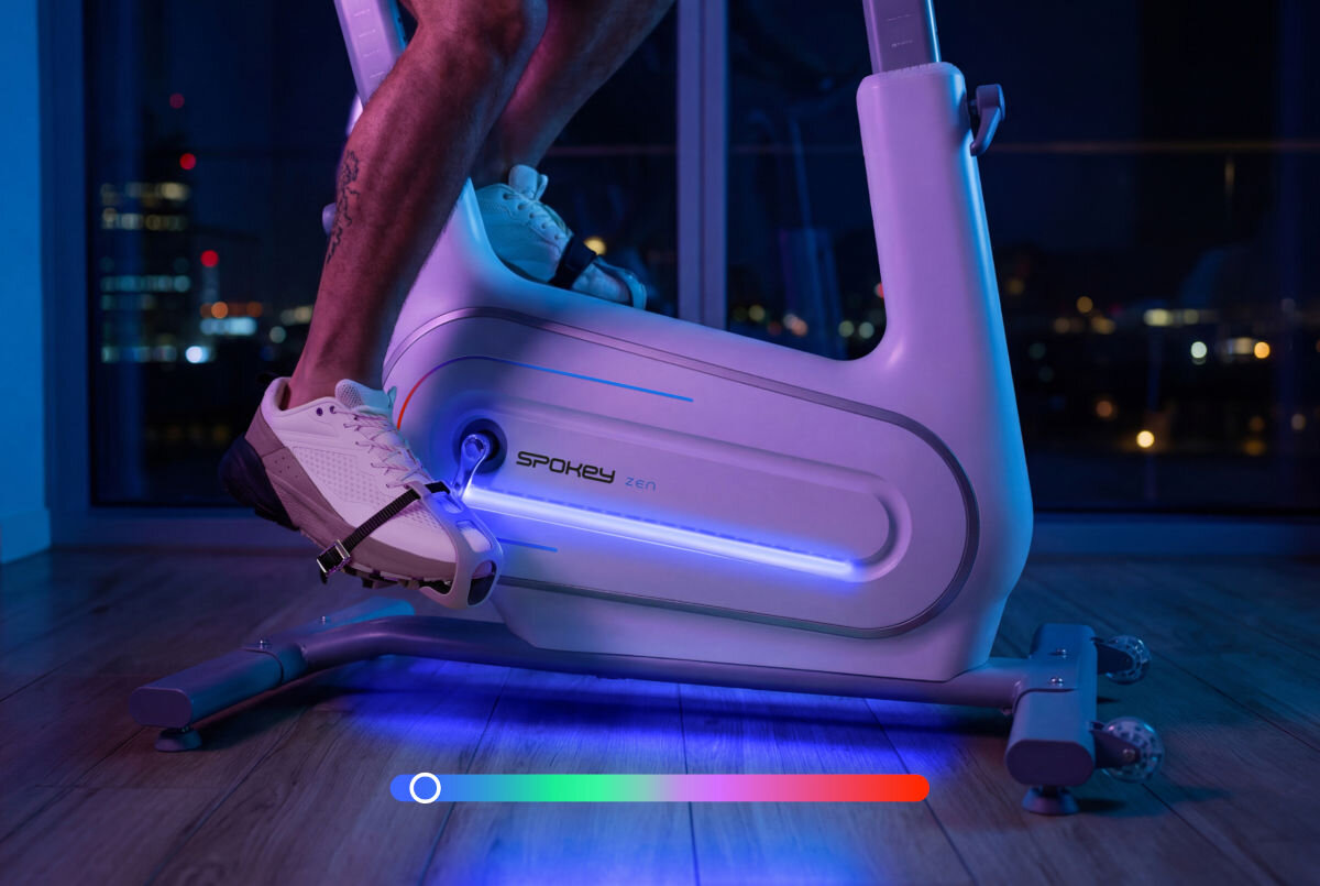 Osoba korzysta z roweru elektromagnetycznego SPOKEY Zen w pomieszczeniu z widokiem na miasto nocą. Na rowerze widoczne jest logo \'Spokey zen\' oraz podświetlenie LED w kolorze niebieskim.