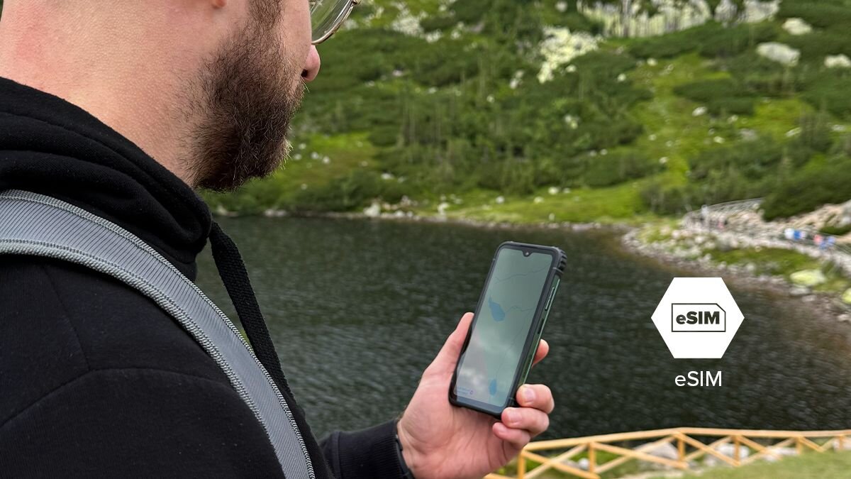 Osoba trzyma smartfon nad jeziorem, w tle widoczny jest zalesiony teren. Obok telefonu znajduje się symbol z napisem \'eSIM\'.