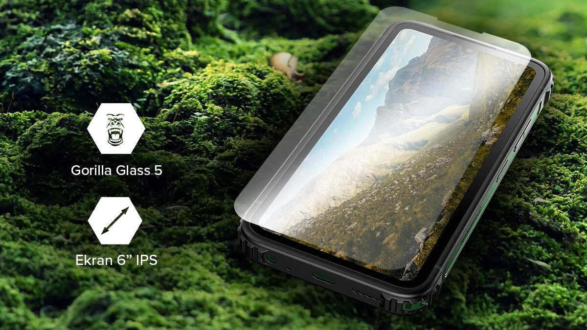 Smartfon z wyświetlaczem pokazującym krajobraz górski, leży na tle zielonego mchu. Obok widoczne są ikony z napisami: \'Gorilla Glass 5\' i \'Ekran 6” IPS\'.