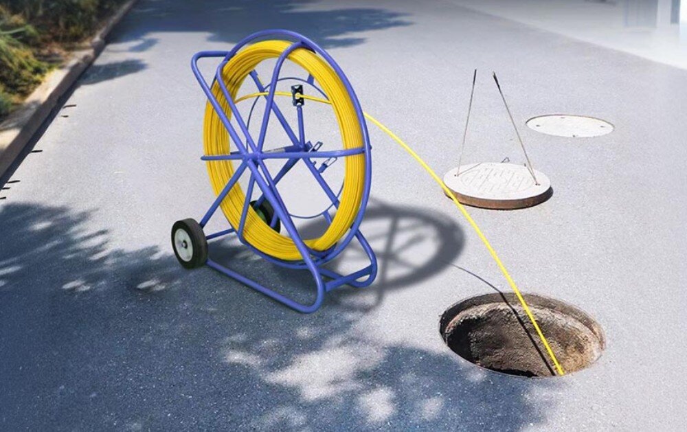 Na obrazie widoczny jest żółty kabel nawinięty na niebieski stojak z kółkami, ustawiony na asfalcie obok otwartego włazu kanalizacyjnego. Kabel jest częściowo rozwinięty i wprowadzony do włazu.
