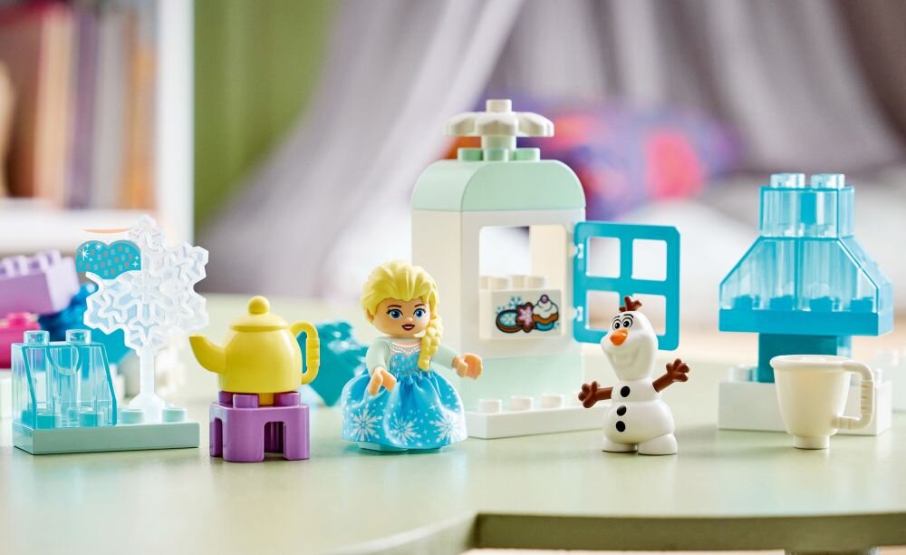 Zestaw LEGO z postaciami Elzy i Olafa oraz akcesoriami, takimi jak dzbanek, kubek i elementy przypominające śnieżynki. W tle widoczne są kolorowe klocki i konstrukcje z przezroczystymi niebieskimi elementami.