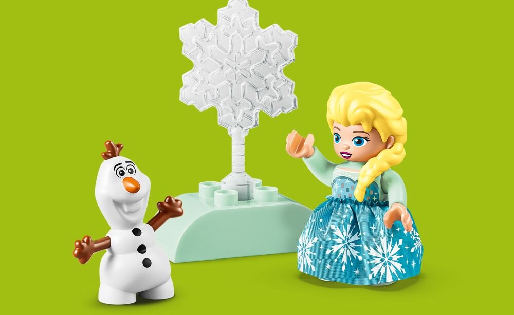 Na zielonym tle znajdują się dwie figurki LEGO: postać z blond włosami w niebieskiej sukience oraz bałwanek z marchewkowym nosem. Obok nich stoi biała śnieżynka na podstawce.