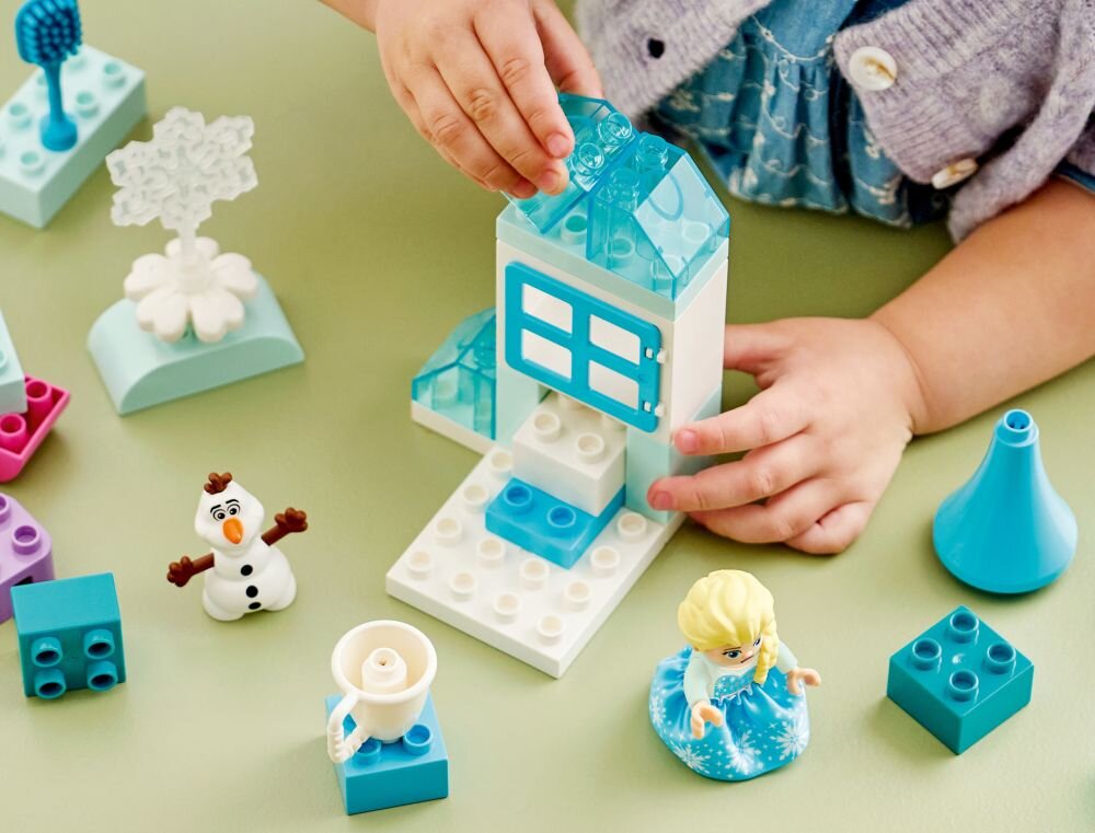 Dziecko bawi się zestawem klocków LEGO z figurkami Elzy i Olafa oraz różnymi elementami konstrukcyjnymi w odcieniach niebieskiego i białego. Na stole znajdują się także inne klocki i akcesoria, takie jak kubek i drzewko.