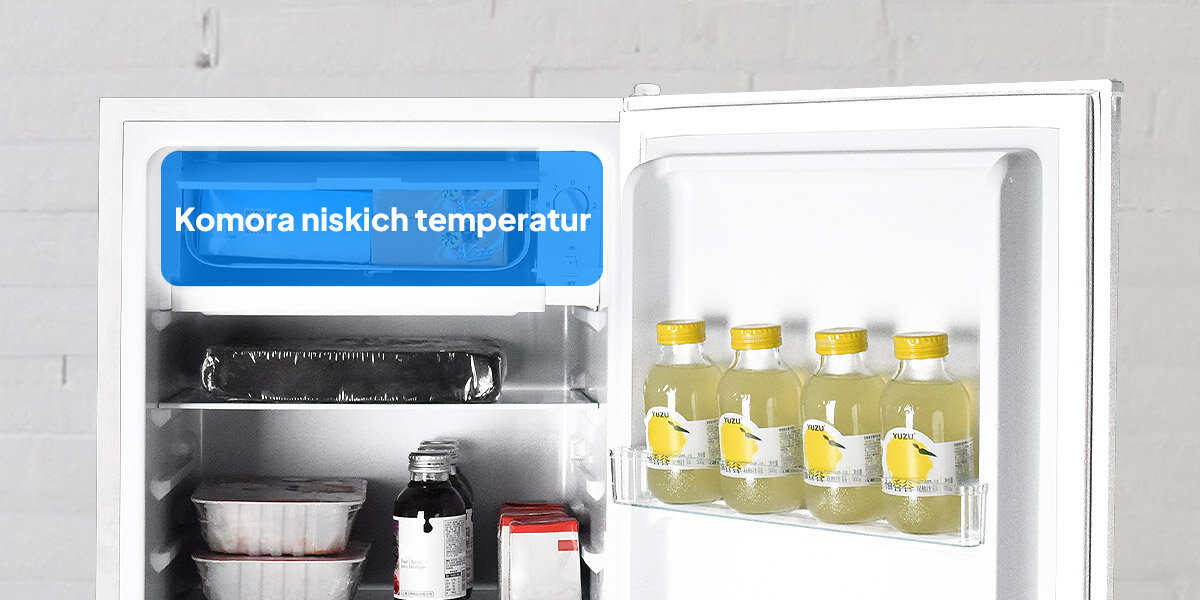 Otwarta lodówka z widocznymi półkami, na których znajdują się różne produkty spożywcze, w tym butelki z napojami na drzwiach. Na górnej półce widoczny jest napis \'Komora niskich temperatur\'.