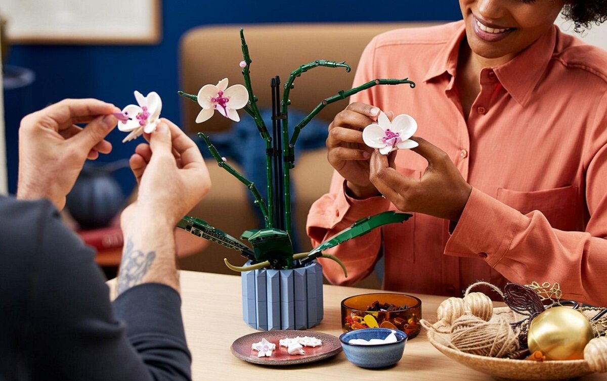 Dwie osoby składają model orchidei z klocków LEGO, który stoi na stole w niebieskiej doniczce. Na stole znajdują się również miseczki z klockami i dekoracyjne elementy.