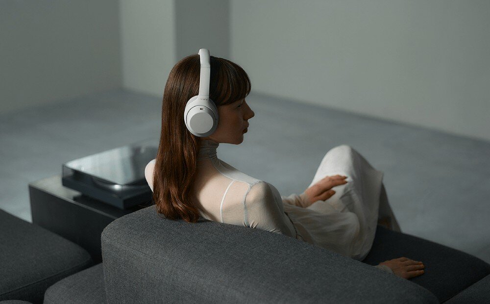 Słuchawki nauszne SONY WH-1000XM4C Kobieta w białej bluzce siedzi na szarej kanapie ze słuchawkami na głowie Patrzy w bok, a za nią znajduje się gramofon
