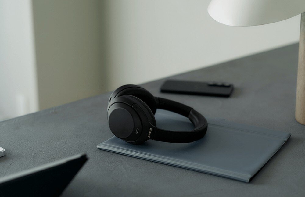 Słuchawki nauszne SONY WH-1000XM4C Słuchawki leżą na zamkniętym etui lub notesie na biurku W tle widać telefon oraz lampkę