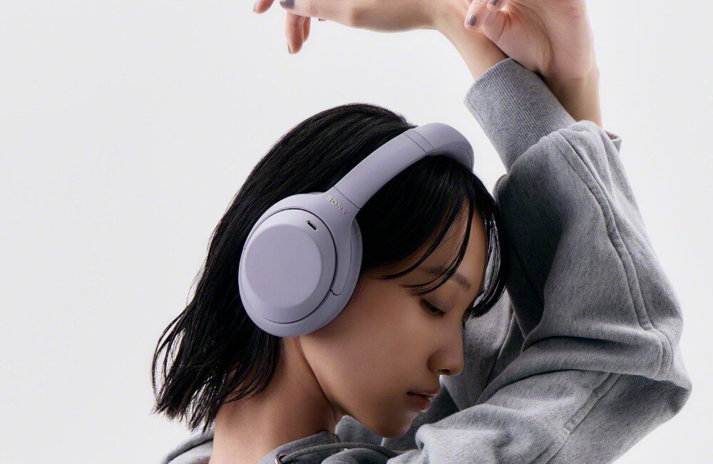 Słuchawki nauszne SONY WH-1000XM4C Kobieta w białej bluzce siedzi na szarej kanapie ze słuchawkami na głowie Patrzy w bok, a za nią znajduje się gramofon
