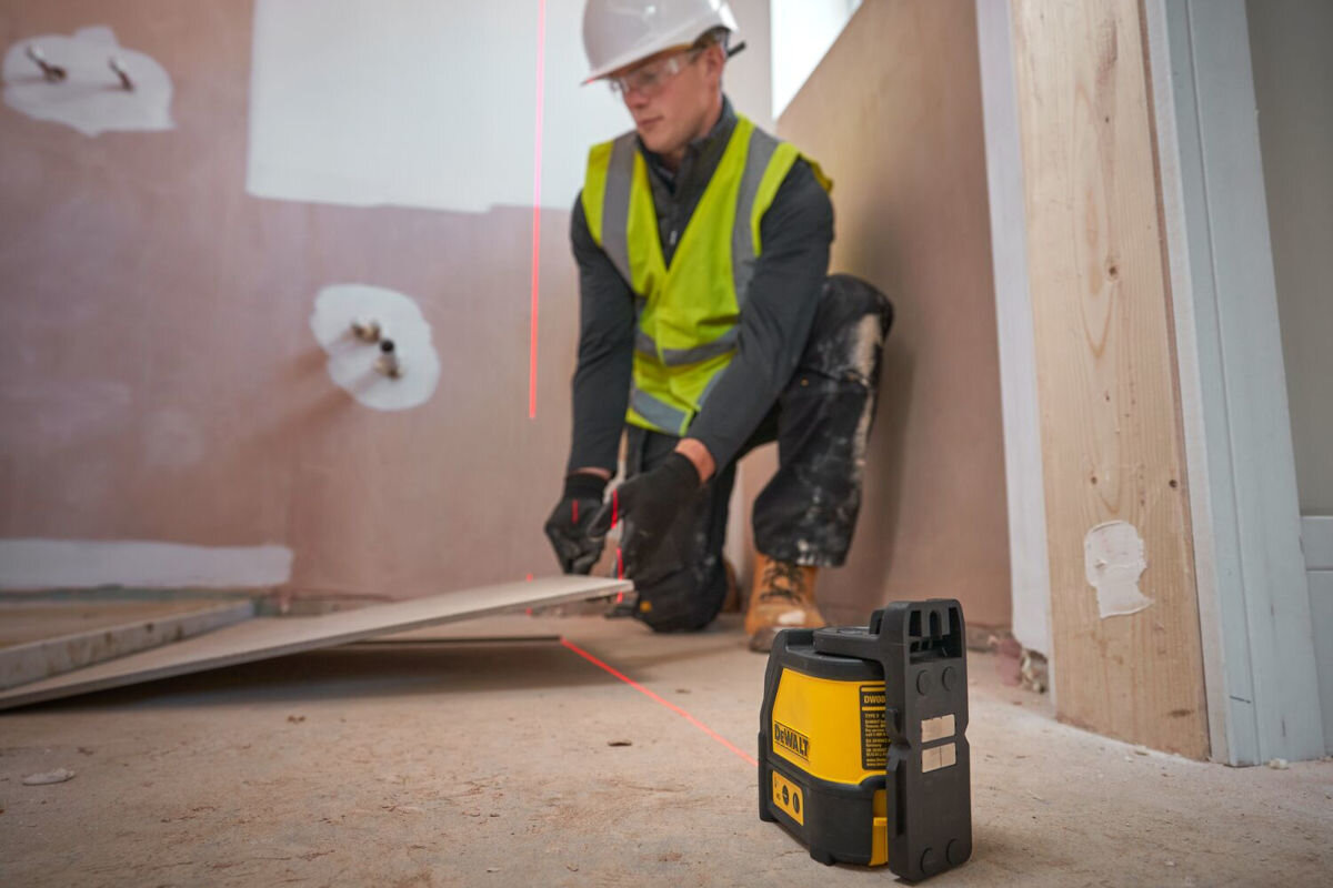 Laser krzyżowy DEWALT DW088K, over-mold, typ, obudowa, uszkodzenia, laser stoi na podłodze, obok pracownik budowy pracuje