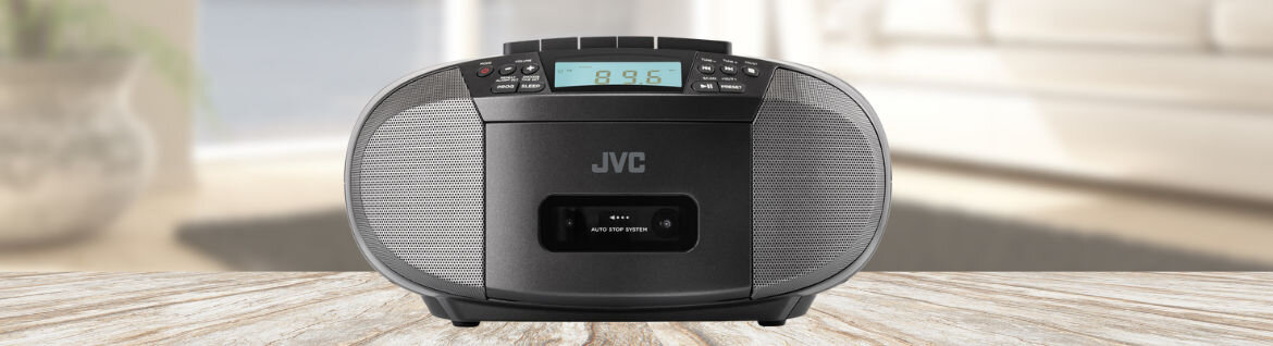 Czarny radioodtwarzacz z wyświetlaczem pokazującym \'89.5\' i logo JVC na przednim panelu. Urządzenie stoi na drewnianej powierzchni w jasnym pomieszczeniu.