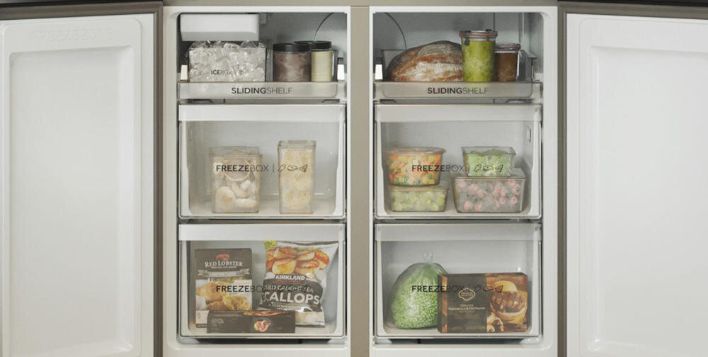 Wnętrze lodówki z widocznymi półkami oznaczonymi jako \'SLIDING SHELF\' i \'FREEZE BOX\'. Na półkach znajdują się różne produkty spożywcze, w tym pojemniki z lodem, słoiki, pieczywo i opakowania z mrożonkami.