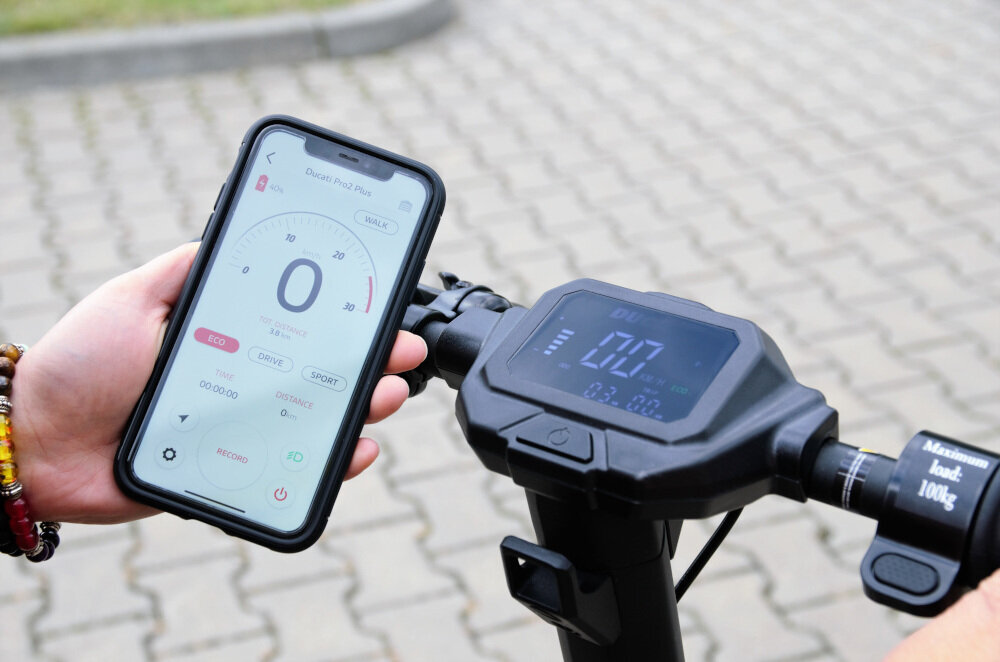 Osoba trzyma smartfon z aplikacją pokazującą prędkość 0 km/h, obok widoczny jest wyświetlacz hulajnogi elektrycznej z podobnym wskazaniem. Na kierownicy znajduje się napis \'Aluminium load. 100kg.\'.