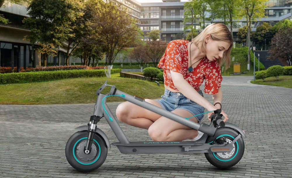 Kobieta siedzi na ziemi, trzymając szarą hulajnogę elektryczną z dużymi kołami i amortyzacją. W tle widać park z drzewami i budynkami.