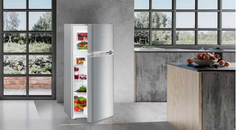 Srebrna lodówka z otwartymi drzwiami, wypełniona różnorodnymi produktami spożywczymi, stoi w nowoczesnej kuchni z dużymi oknami. Na blacie kuchennym znajduje się misa z owocami.