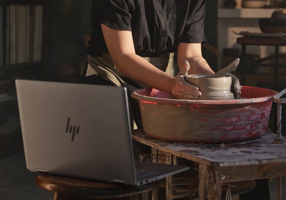 Osoba formuje gliniane naczynie na kole garncarskim, obok stoi otwarty laptop z widocznym logo HP.