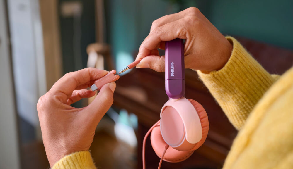 Osoba trzyma różowo-fioletowe słuchawki nauszne z widocznym logo \'Philips\' oraz wtyczkę audio. W tle znajduje się rozmyte wnętrze pomieszczenia.
