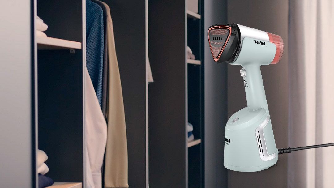 Na pierwszym planie widoczna jest parownica do ubrań z logo \'Tefal\', ustawiona na tle otwartej szafy z ubraniami. Parownica ma biało-czerwony kolor i jest podłączona do prądu.