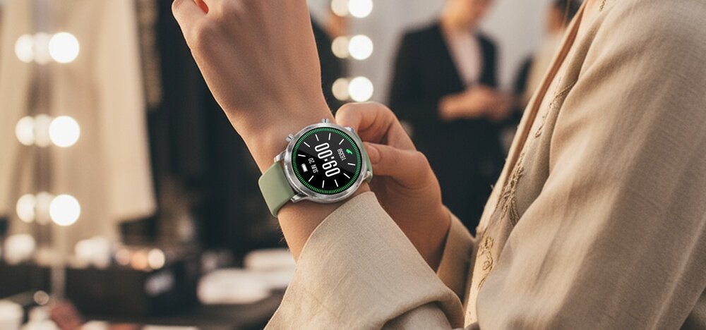 Osoba nosi smartwatch z zielonym paskiem i srebrną kopertą. Na wyświetlaczu widoczny jest czas 10:08:00.