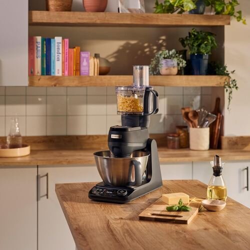 Na blacie kuchennym stoi robot kuchenny z metalową misą i przystawką do siekania. W tle widać drewniane półki z książkami i roślinami.