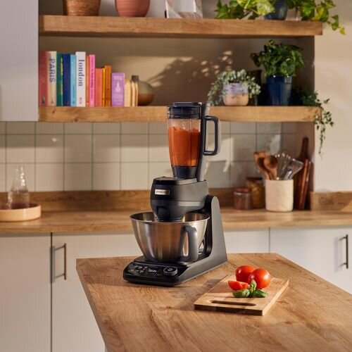 Na blacie kuchennym stoi robot kuchenny z metalową misą i blenderem z czerwonym płynem. W tle widoczne są drewniane półki z książkami i roślinami.