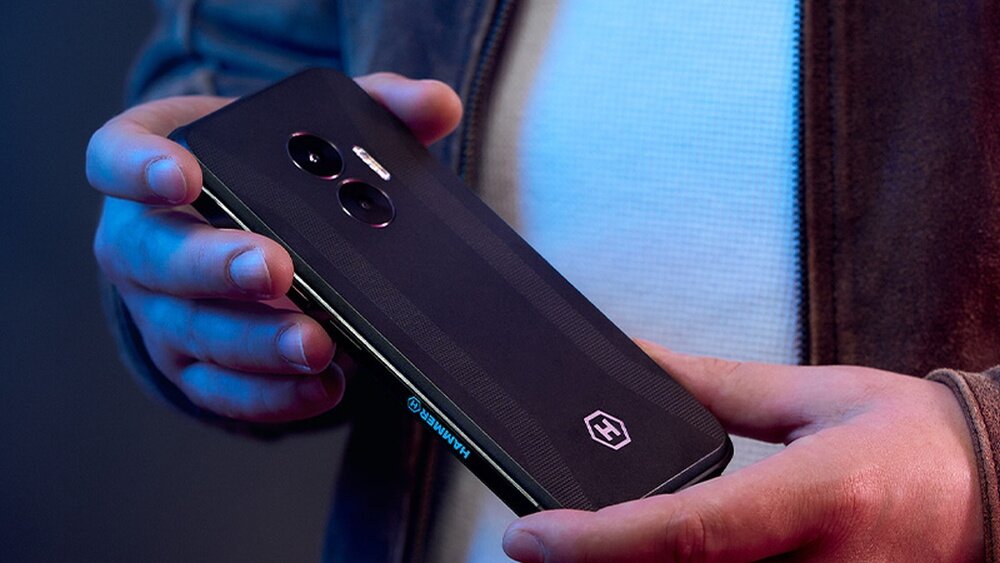 Osoba trzyma czarny smartfon z dwoma aparatami i logo HAMMER na obudowie. Telefon ma wyraźnie widoczne przyciski boczne.
