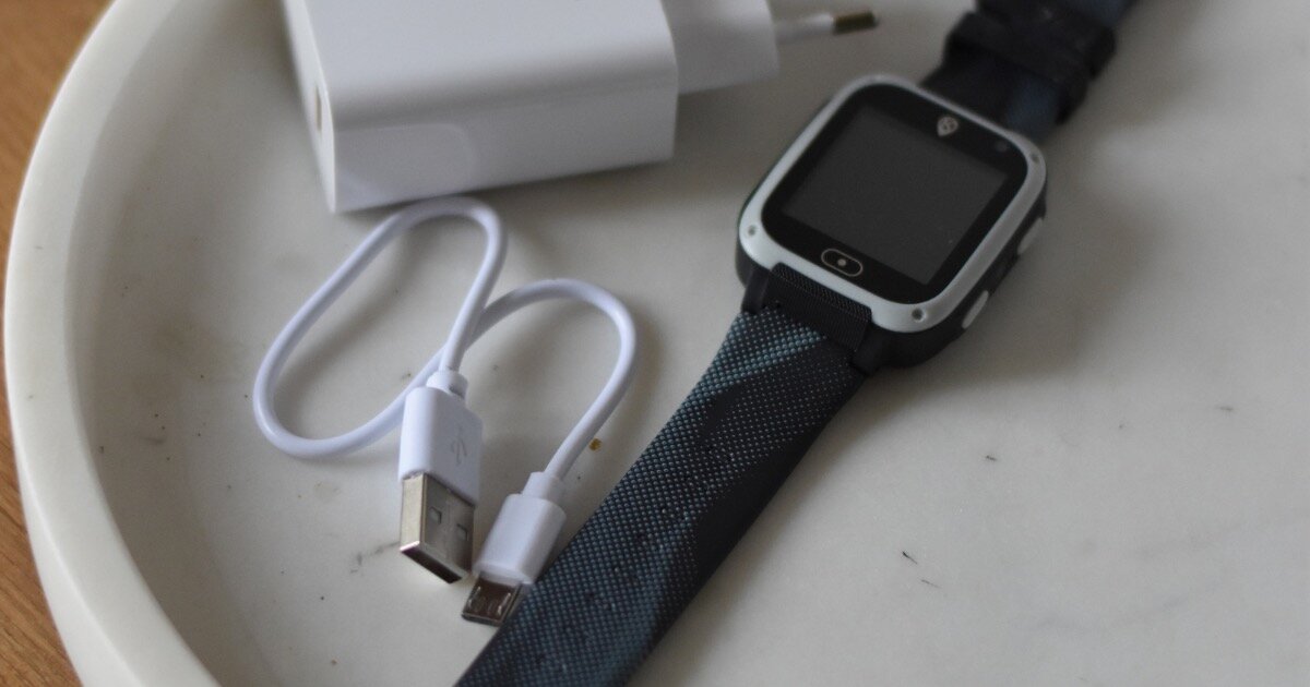 Na białym talerzu leży czarny smartwatch z prostokątnym ekranem, obok znajduje się biały kabel USB oraz ładowarka.