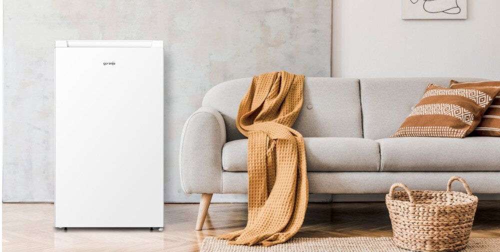 Biała lodówka z logo \'gorenje\' stoi obok szarej kanapy z brązowym kocem. Na podłodze leży pleciony kosz i dywanik.