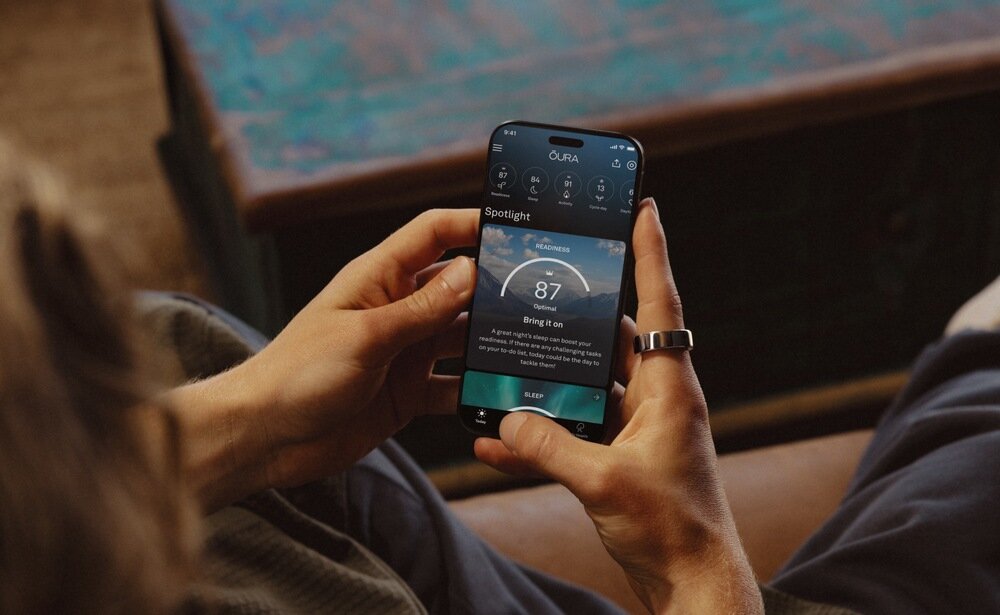 Osoba trzyma smartfon z aplikacją OURA, na ekranie widoczny jest wynik \'87\' oraz tekst \'Bring it on\'. Na palcu osoby znajduje się srebrny pierścień.