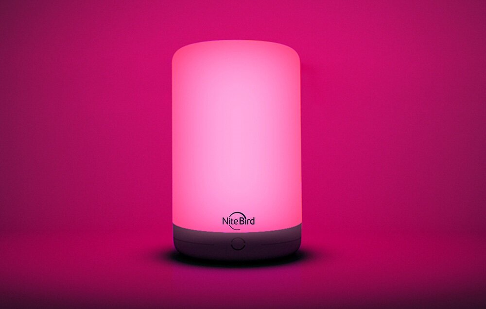 Na obrazie widoczna jest inteligentna lampka nocna emitująca różowe światło. Na podstawie lampki znajduje się logo \'NiteBird\'.