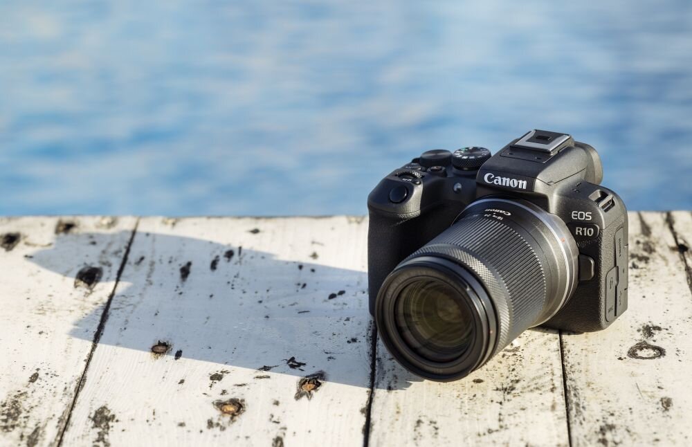 Aparat bezlusterkowy z obiektywem leży na drewnianej powierzchni. Na korpusie widoczne są napisy \'Canon\' i \'EOS R10\'.