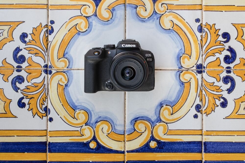 Aparat fotograficzny z logo \'Canon\' i oznaczeniem \'EOS R10\' umieszczony na tle ozdobnych płytek ceramicznych w kolorach niebieskim, żółtym i białym.