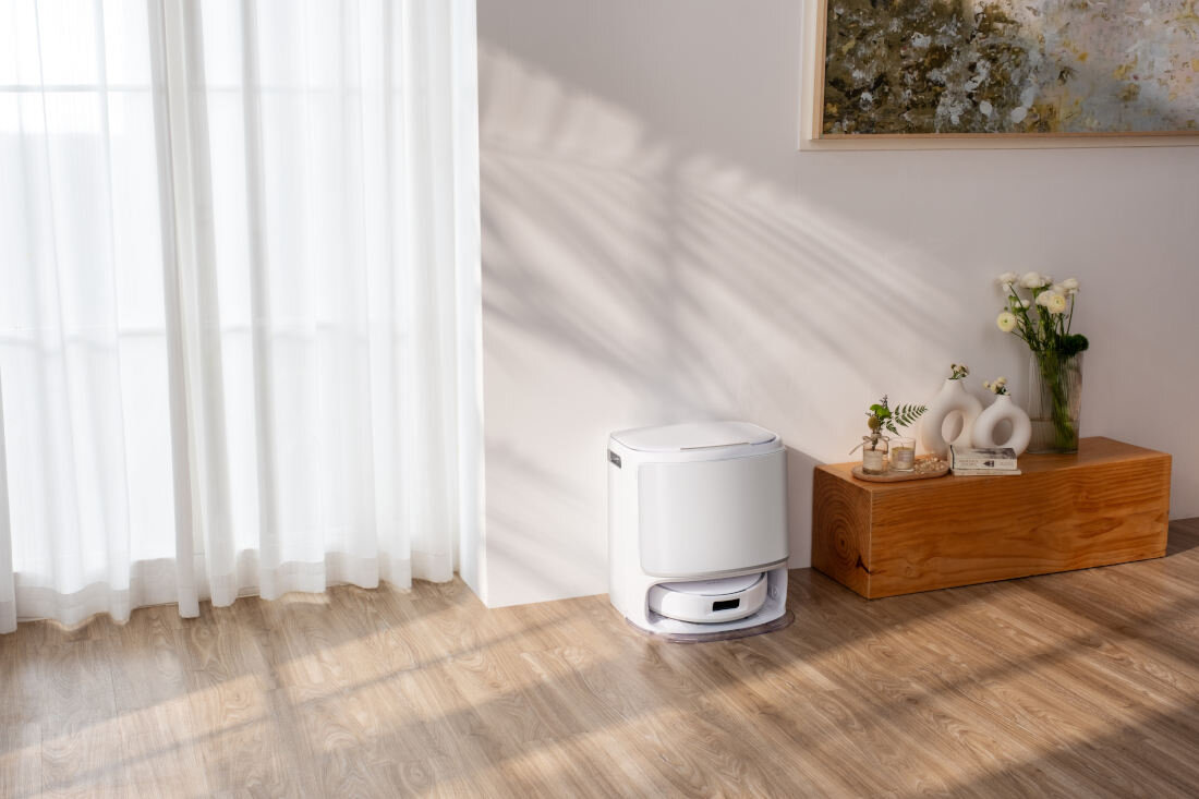 Na drewnianej podłodze stoi biały robot sprzątający z bazą ładującą. Obok znajduje się drewniana ławka z wazonem i dekoracjami, a w tle widać zasłony i obraz na ścianie.