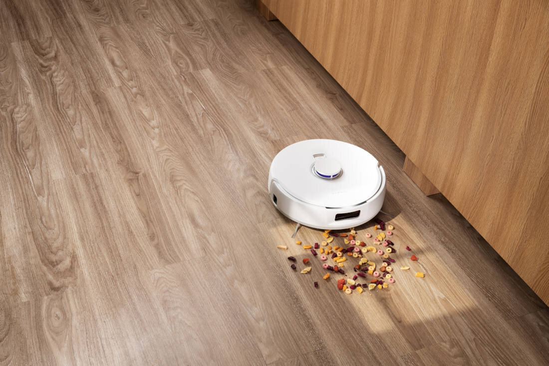 Biały robot sprzątający znajduje się na drewnianej podłodze obok rozsypanych płatków śniadaniowych. Robot jest w trakcie sprzątania, a w tle widoczna jest drewniana szafka.