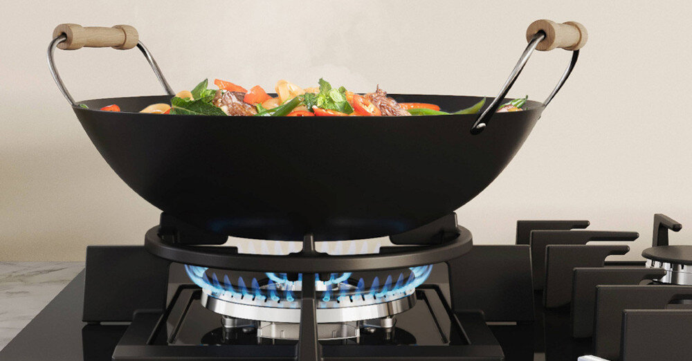 Czarna patelnia wok z drewnianymi uchwytami stoi na palniku gazowym, na którym widać niebieski płomień. W patelni znajdują się kolorowe warzywa i kawałki mięsa.