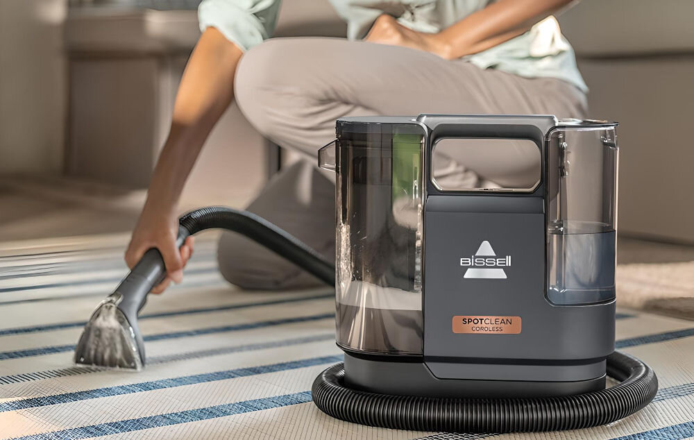 Na obrazie widoczny jest odkurzacz piorący z logo \'Bissell\' i napisem \'SpotClean Cordless\'. W tle osoba używa urządzenia do czyszczenia dywanu.