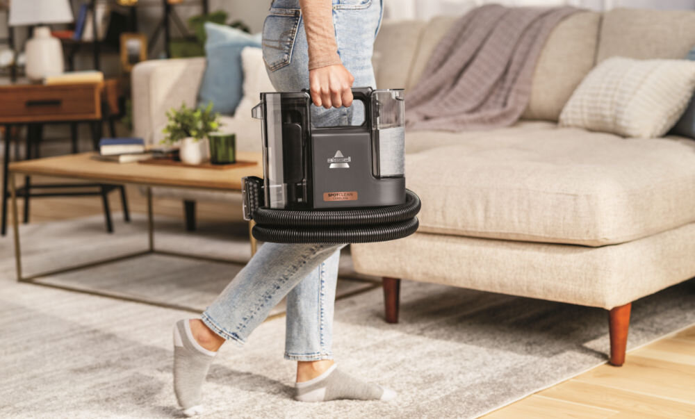 Osoba trzyma odkurzacz piorący BISSELL SpotClean Cordless 3681N w ręce, stojąc w salonie z sofą i stolikiem w tle. Na urządzeniu widoczne jest logo producenta.