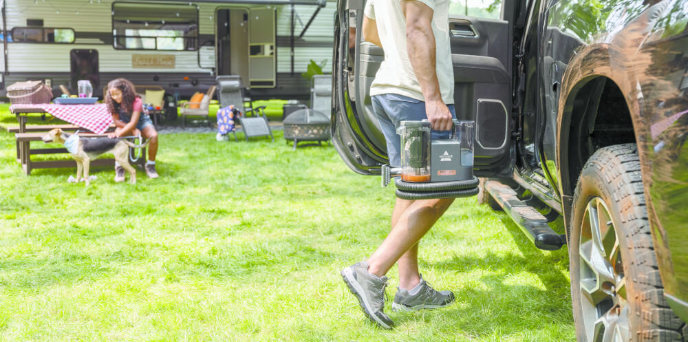 Osoba wychodzi z samochodu, trzymając odkurzacz piorący BISSELL SpotClean Cordless 3681N. W tle widać przyczepę kempingową i osobę siedzącą przy stole piknikowym z psem.