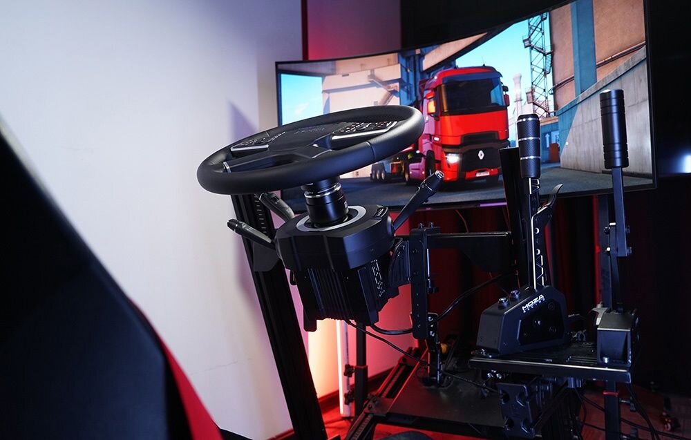 Na zdjęciu znajduje się symulator jazdy z kierownicą i pedałami, przed dużym ekranem wyświetlającym obraz ciężarówki na drodze. W tle widoczne są elementy konstrukcji symulatora oraz oświetlenie.