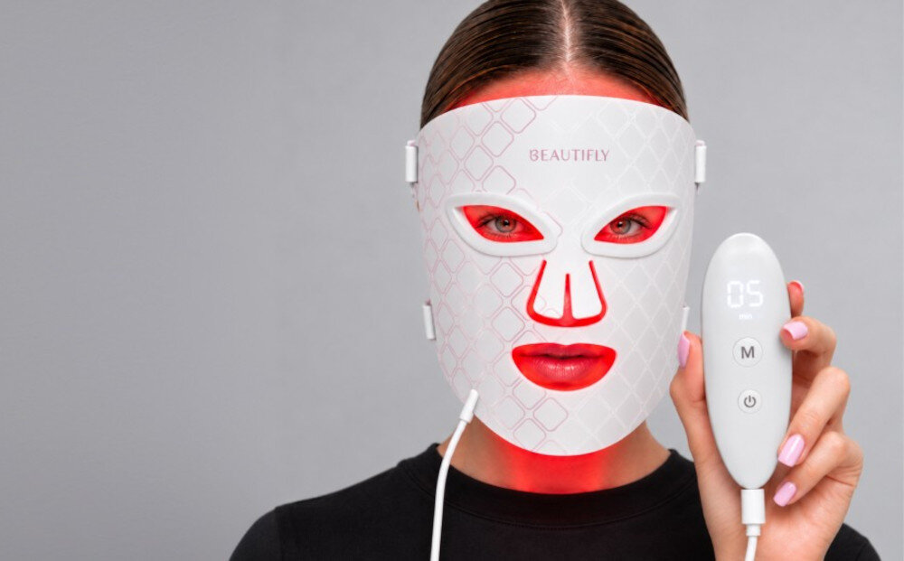 Osoba nosi białą maskę LED z napisem \'BEAUTIFLY\', która emituje czerwone światło. Obok trzyma urządzenie z wyświetlaczem pokazującym \'05\' i dwoma przyciskami oznaczonymi literami \'M\' i \'C\'.