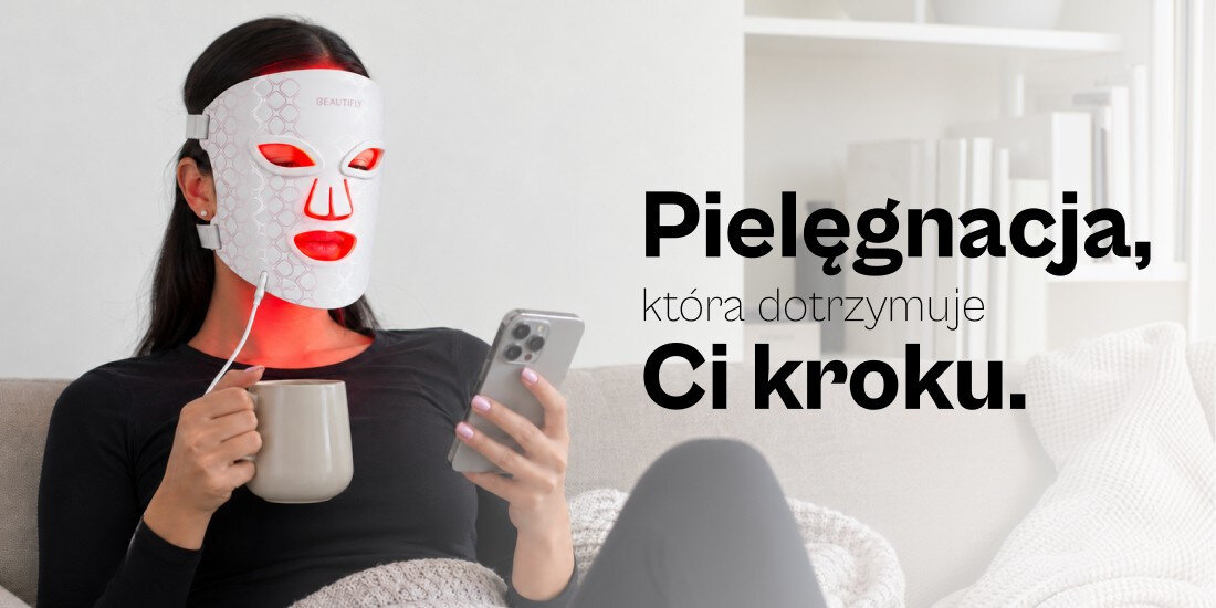 Osoba siedzi na kanapie, nosząc białą maskę LED na twarzy i trzymając kubek oraz smartfon. Obok widoczny jest tekst: \'Pielęgnacja, która dotrzymuje Ci kroku.\'