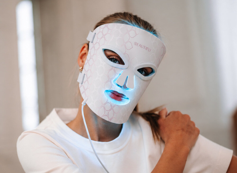 Osoba nosi białą maskę LED do twarzy z napisem \'BEAUTIFLY\', która emituje niebieskie światło wokół oczu i ust. Maska jest przymocowana paskami i podłączona kablem.